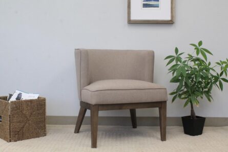 A Ava Accent Chair-Beige sits next to a green plant and a wicker basket with magazines, against a wall with a framed picture.