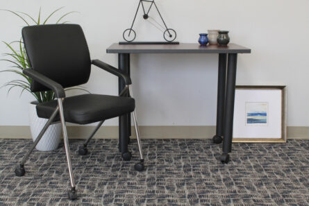 A modern workspace with a TOS Black Caressoft Plus Training Chair With Chrome Frame, metal frame desk, decorative sculptures, a small blue pot, and a framed picture leaning against the wall on a carpeted floor.