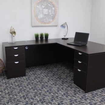 A modern office with a dark brown l-shaped desk containing a laptop, decorative plants, a lamp, and art pieces, set against a gray carpeted floor.