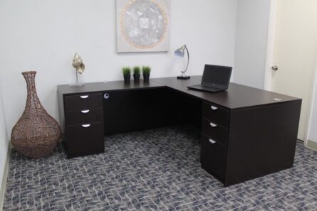 A modern office with a dark brown l-shaped desk containing a laptop, decorative plants, a lamp, and art pieces, set against a gray carpeted floor.