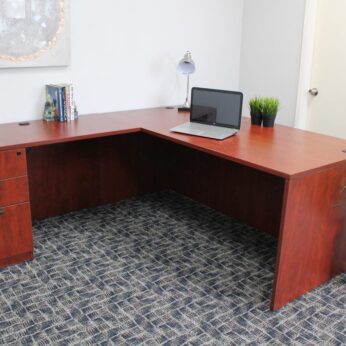 An Laminate 71 Inch Executive L-Shape Corner Desk with File Storage Pedestal, Cherry with a laptop, lamp, and books in an office room with gray carpet and decorative plants.