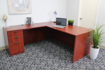 An Laminate 71 Inch Executive L-Shape Corner Desk with File Storage Pedestal, Cherry with a laptop, lamp, and books in an office room with gray carpet and decorative plants.