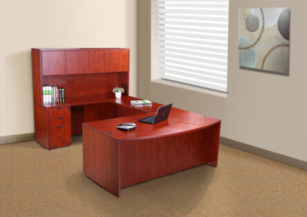 Modern office setup with an Executive U-Shaped Curved Bow Desk with File Storage Pedestal and Hutch, Cherry, laptop, and shelving unit in a room with beige walls and a window with blinds.