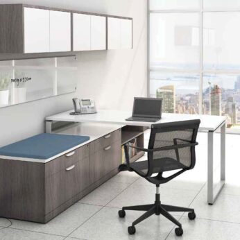 Modern office with an Elements Series L shaped desk #4, chair, and laptop, overlooking a cityscape through large windows. a fan and shelving unit are also visible.