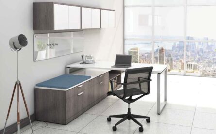 Modern office with an Elements Series L shaped desk #4, chair, and laptop, overlooking a cityscape through large windows. a fan and shelving unit are also visible.