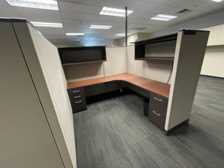 Empty Haworth 67" tall cubicle with a corner desk, overhead cabinets, and two drawer units in a room with gray carpeting.