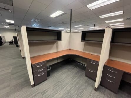 An empty office space with two Haworth 67" tall cubicles featuring corner desks, cabinets, and overhead storage units under fluorescent lighting.