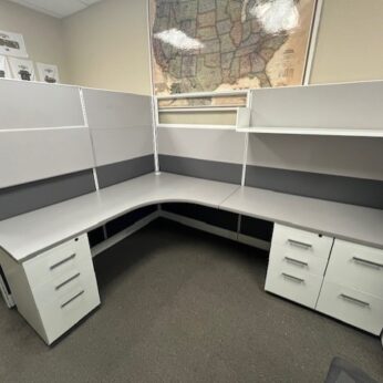 An empty office cubicle with two Friant Novo Series desks forming an l-shape, white filing cabinets, and grey partition walls, with a world map on the wall.