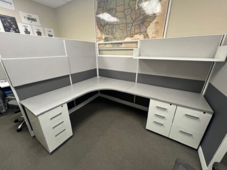 An empty office cubicle with two Friant Novo Series desks forming an l-shape, white filing cabinets, and grey partition walls, with a world map on the wall.